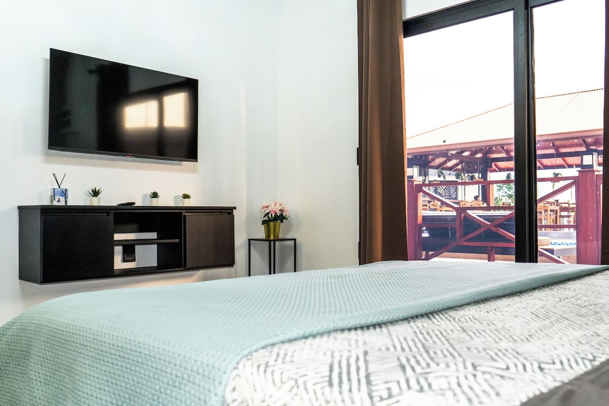 Ein Hotelzimmer mit einem Bett, einem an der Wand montierten Fernseher, einem kleinen Tisch mit Blumen in einer Vase und Blick auf einen Außenbereich durch ein großes Fenster.