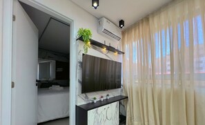 Una habitación de hotel moderna con un televisor de pantalla plana grande montado sobre una consola de mármol, una pequeña planta en maceta y una ventana con cortinas traslúcidas.
