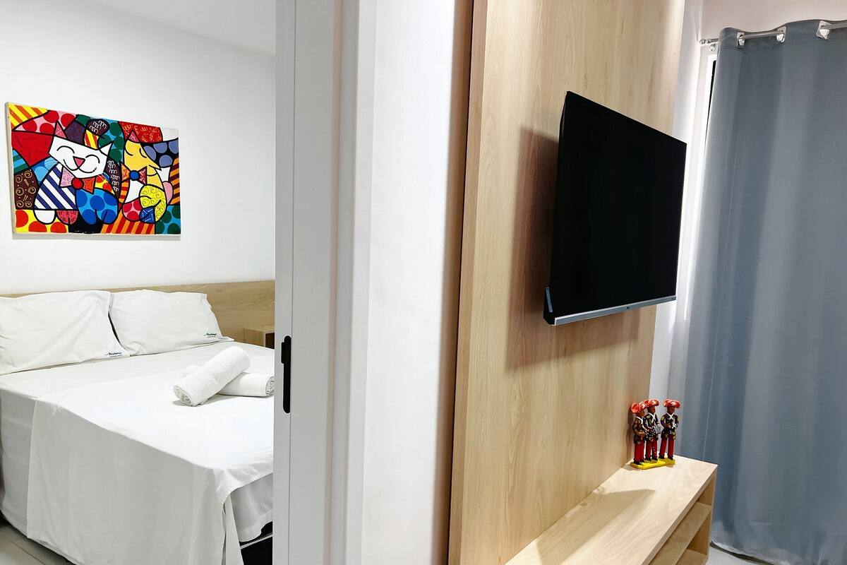 Une chambre d’hôtel avec un lit, un téléviseur à écran plat fixé sur un panneau en bois, une œuvre d’art colorée au mur et une petite étagère avec des figurines.