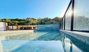 Una piscina con agua cristalina, rodeada por un edificio moderno con amplias ventanas y un muro de piedra.