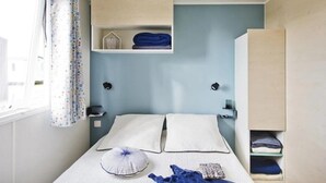 A bedroom with a bed, pillows, a bedside table, a shelf with folded towels, and a window with curtains.