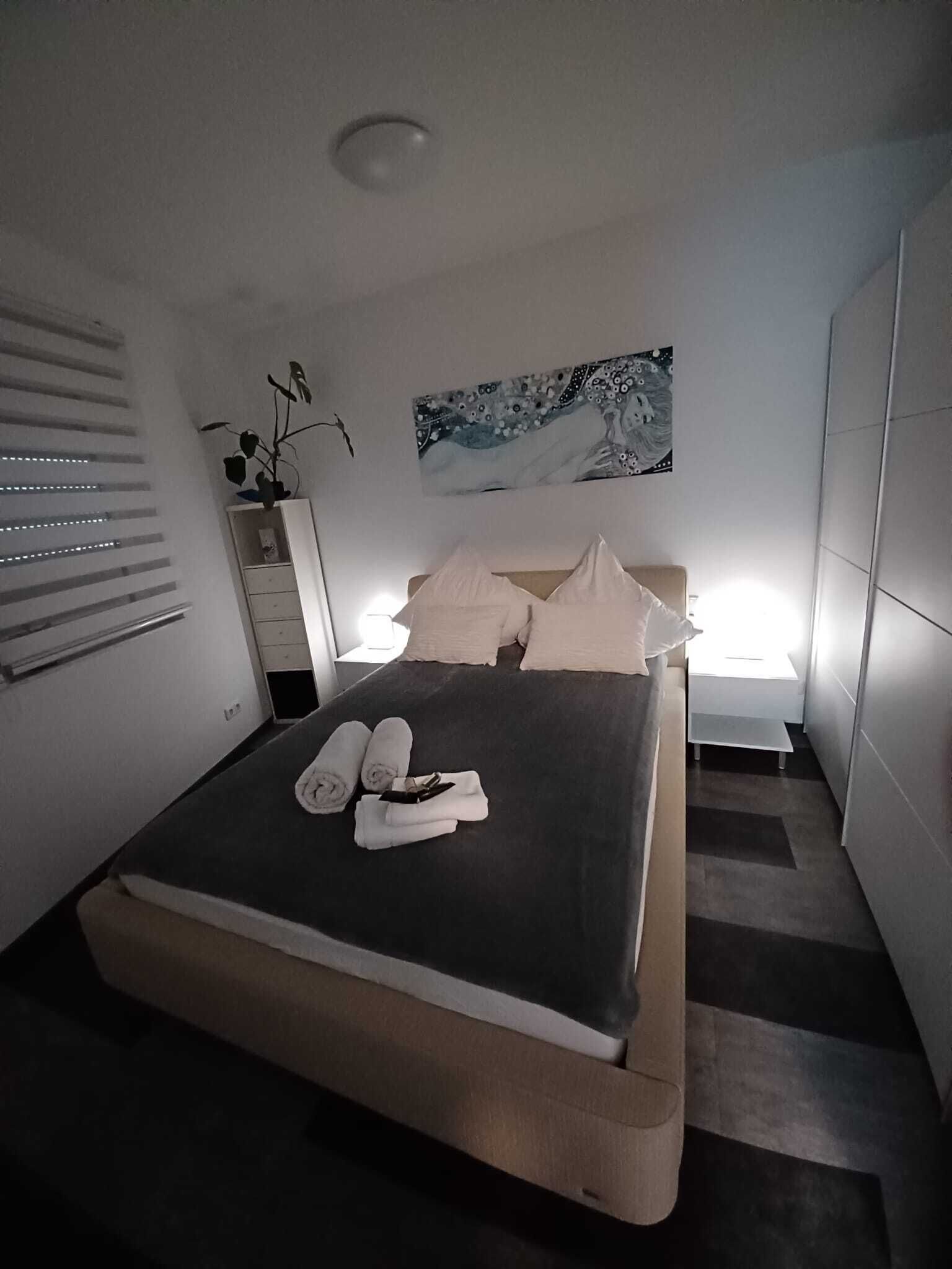 Un dormitorio moderno con cama, mesitas de noche, un cuadro en la pared y una ventana con persianas.