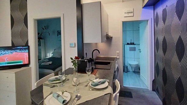 A modern dining area with a round table set for four, a television displaying a tennis match, and a kitchen area visible through an open door.