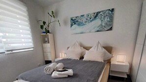 A modern bedroom with a large bed, bedside tables, a nightstand, a wall-mounted painting, and a window with blinds.