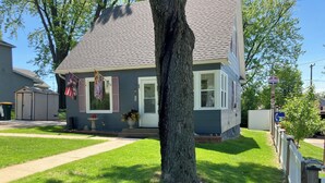 Une maison à deux étages avec un toit gris et des bordures blanches, une pelouse devant la maison avec un arbre et une boîte aux lettres.