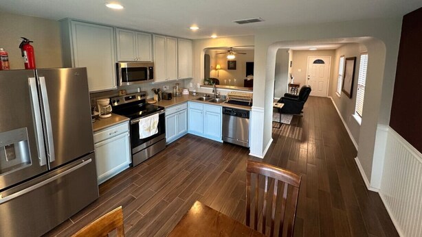 A modern kitchen with stainless steel appliances, a dining area with a wooden table, and a hallway leading to a living space.