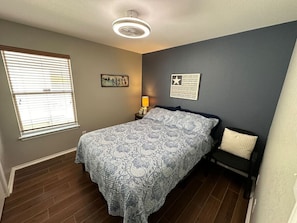 Une chambre à coucher comprenant un lit, une table de chevet, une chaise, une fenêtre avec des stores et un mur orné d’un tableau encadré.