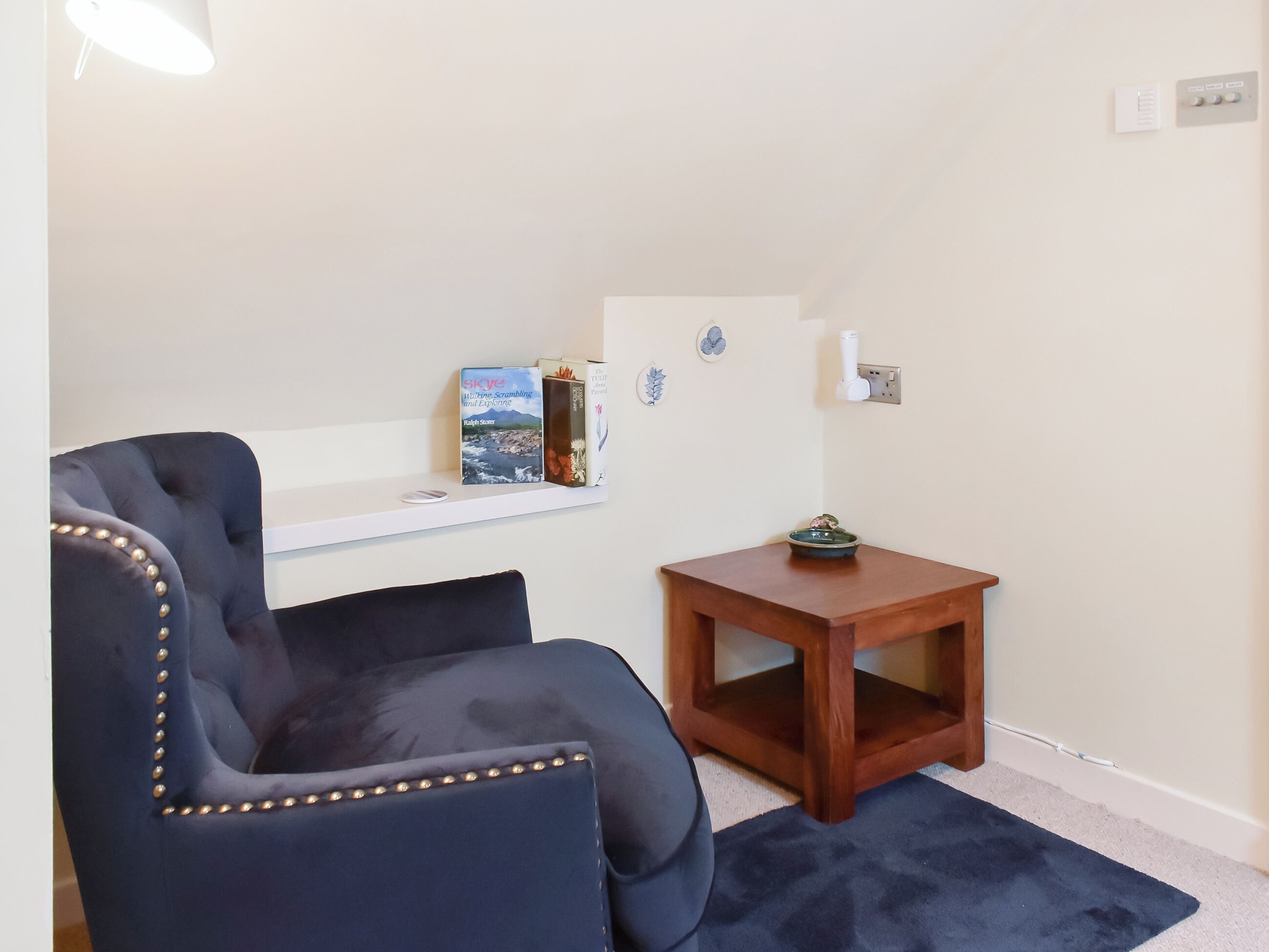 Une pièce comprenant un fauteuil bleu foncé recouvert de tissu, une table d’appoint en bois, une étagère blanche avec des livres et une petite assiette décorative.