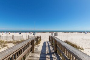 Ein hölzerner Steg führt zu einem Sandstrand bei blauem Himmel.
