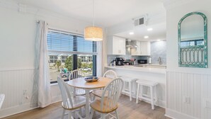 A dining area with a round table and white chairs, a kitchen with white cabinets, and a view of the outside through large windows.