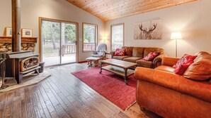 A cozy living room with a stone fireplace, wooden flooring, a red rug, a leather sofa, and a large window offering a view of the outdoors. - Home w/ Deck + Access to Tahoe Donner Amenities! (Truckee)