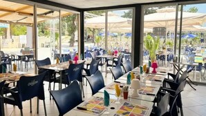 Un espacio exterior con mesas y sillas, ventanas amplias y sombrillas.