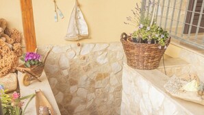 Zona esterna con pareti in pietra, una panca di legno, un cesto di vimini con piante e una piccola carriola piena di fiori.