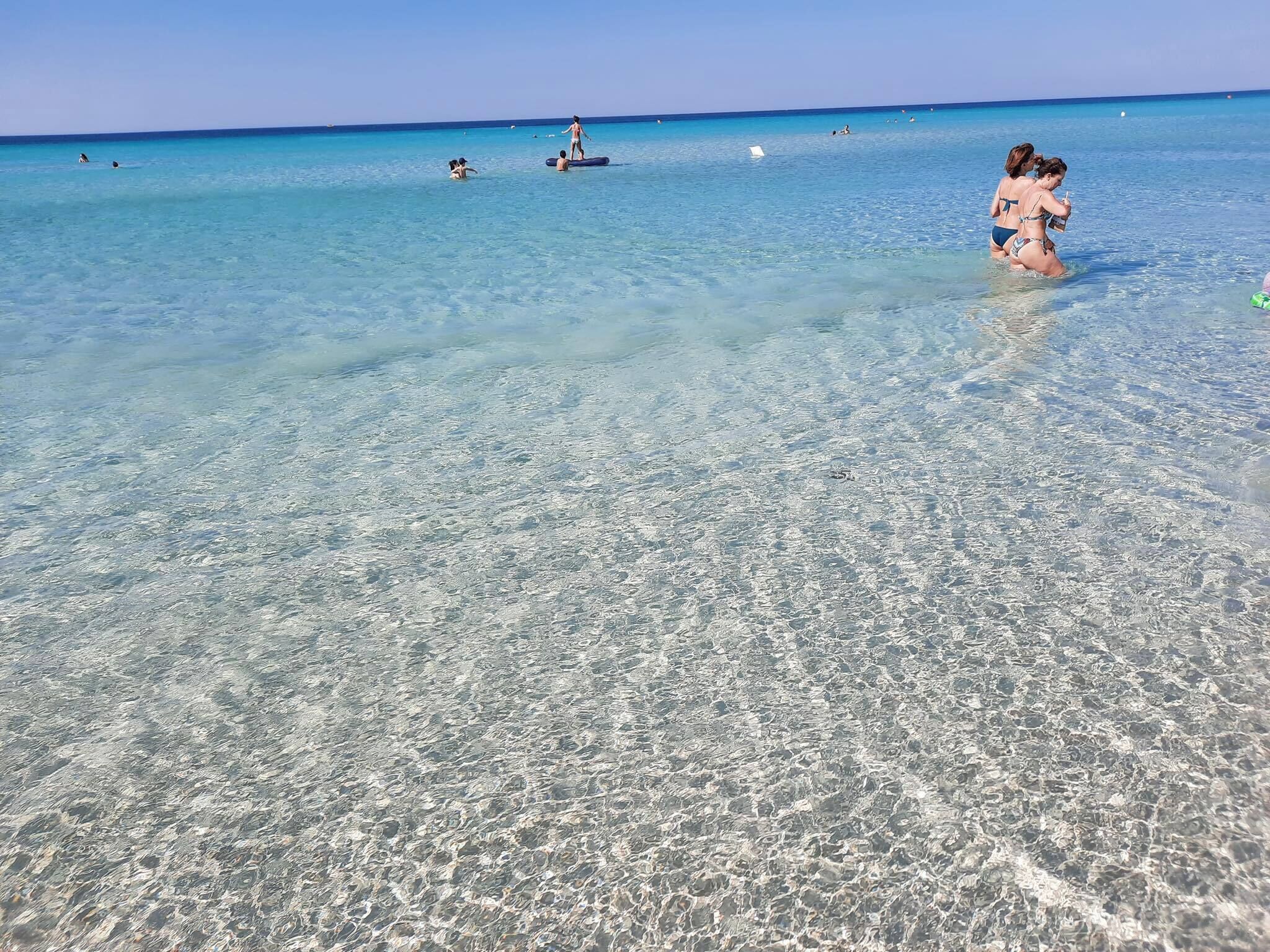 A beach with clear shallow water, people swimming and standing in the water, and a distant horizon.