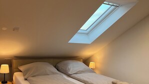 A bedroom with a skylight, a bed with pillows, a nightstand with a lamp, and a wooden floor. - Apartment mit Parkblick, Stilvolle Einrichtung, 1,1km vom Hauptbahnhof Entfernt (Bremerhaven)