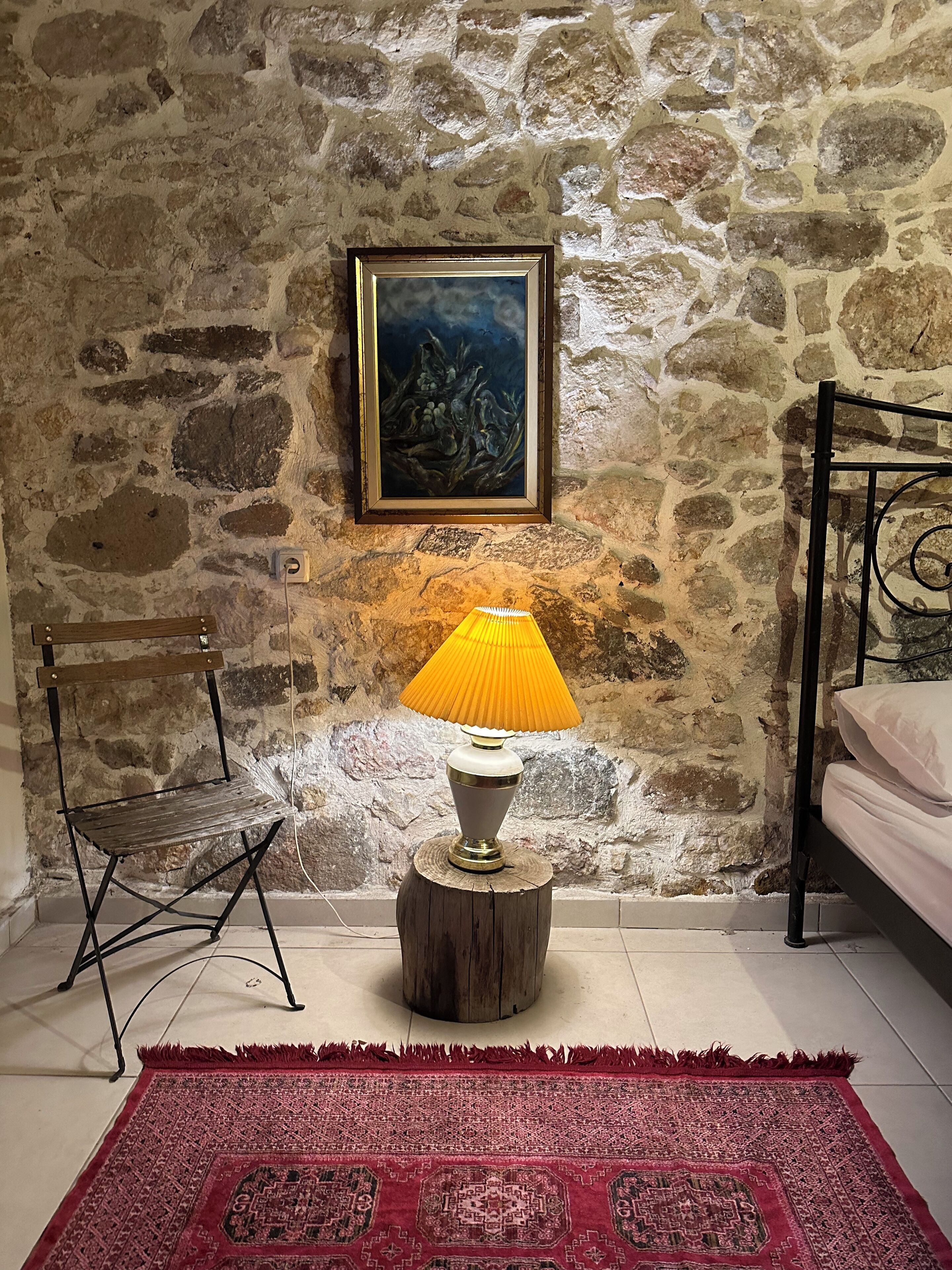 Una habitación con pared de piedra, cama, silla, lámpara y alfombra.