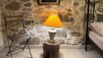 Una habitación con pared de piedra, cama, silla, lámpara y alfombra.