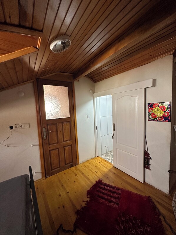 A room with wooden ceiling beams, a wooden door, a white wall, a red rug, and a small window with a decorative pattern. - Sera Pansiyon Ayvalık (Ayvalik)