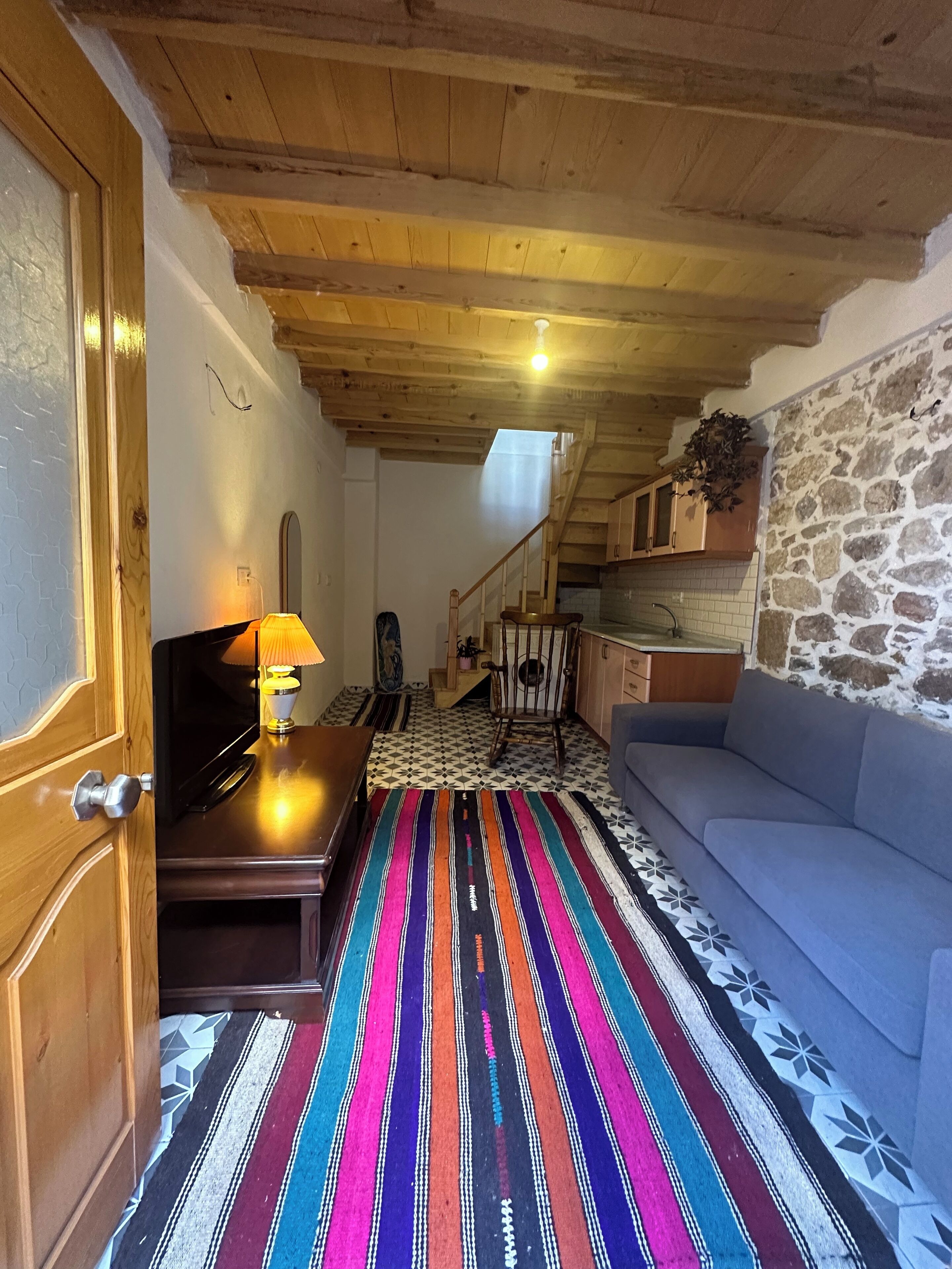 A cozy living room with a wooden ceiling, a colorful striped rug, a sofa, and a television on a wooden table.