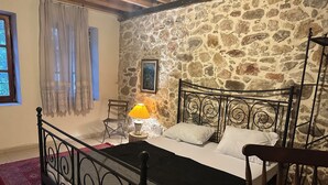 Un dormitorio con pared de piedra, estructura de cama de metal, una silla de madera, una mesita de noche con lámpara y una ventana con cortinas.