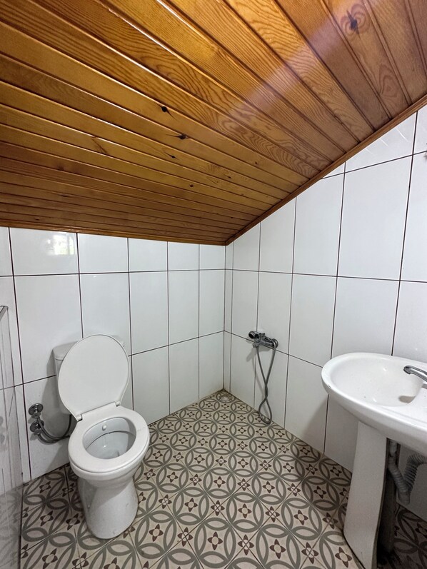 A bathroom with a wooden ceiling, white tiled walls, a patterned floor, a toilet, a shower, and a sink. - Sera Pansiyon Ayvalık (Ayvalik)