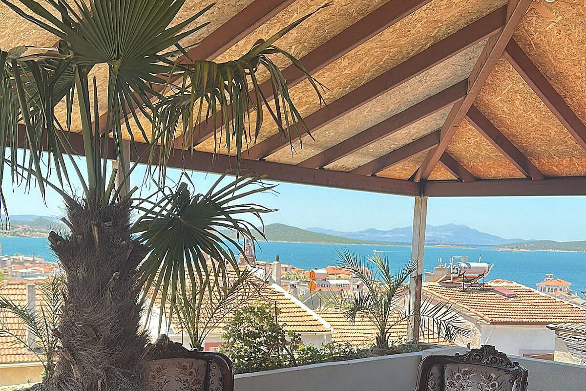 Un espacio de estar al aire libre cubierto con mesa y sillas, una planta en maceta y vistas al mar y edificios.