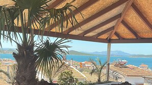 Un espacio de estar al aire libre cubierto con mesa y sillas, una planta en maceta y vistas al mar y edificios.