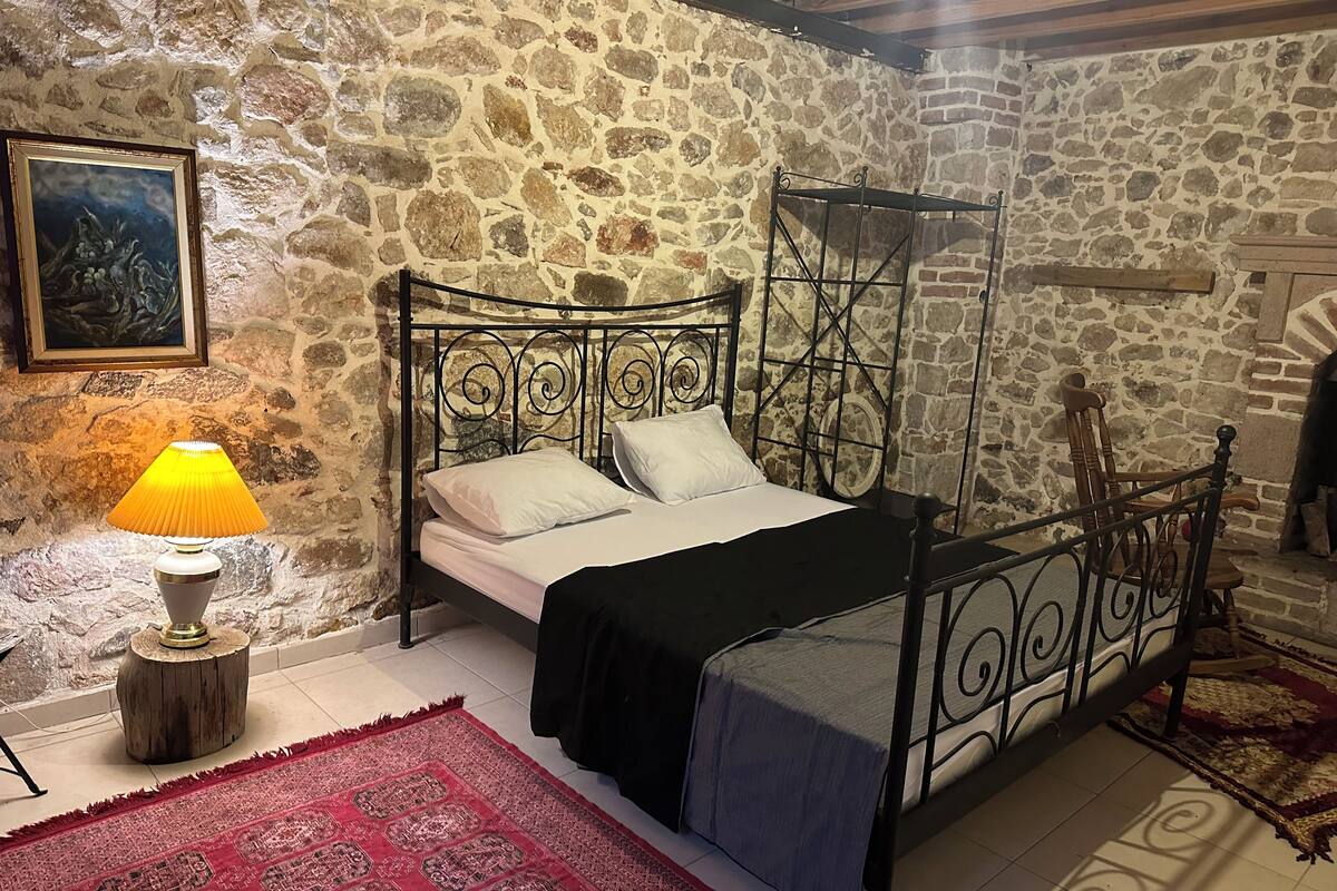 Un dormitorio de paredes de piedra con cama con dosel, una silla de madera y una mesita de noche con lámpara.