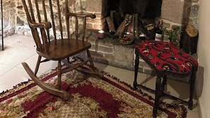Una habitación de paredes de piedra con chimenea, un sillón mecedor y una pequeña mesa con mantel estampado.