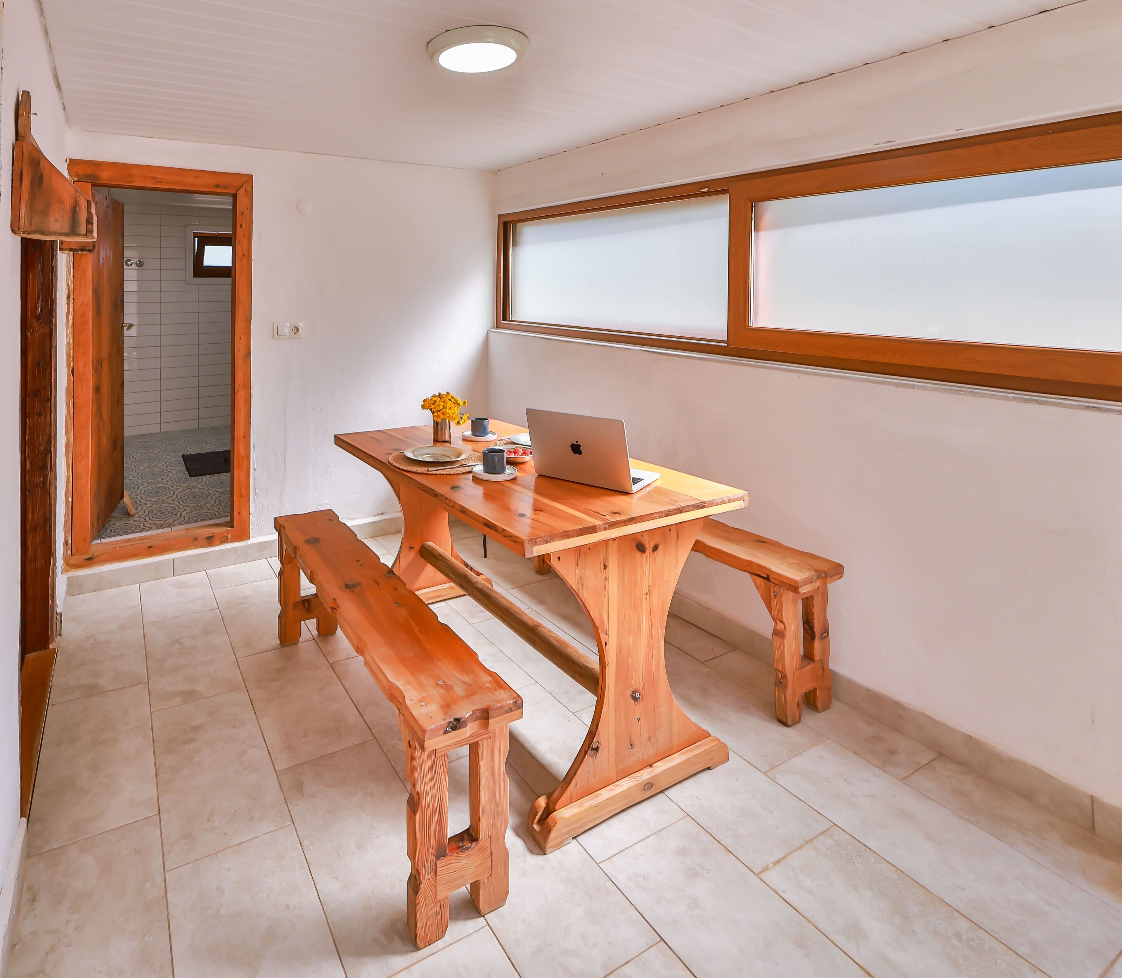 Une table de salle à manger en bois, sur laquelle se trouvent un ordinateur portable, un vase avec des fleurs et un banc.