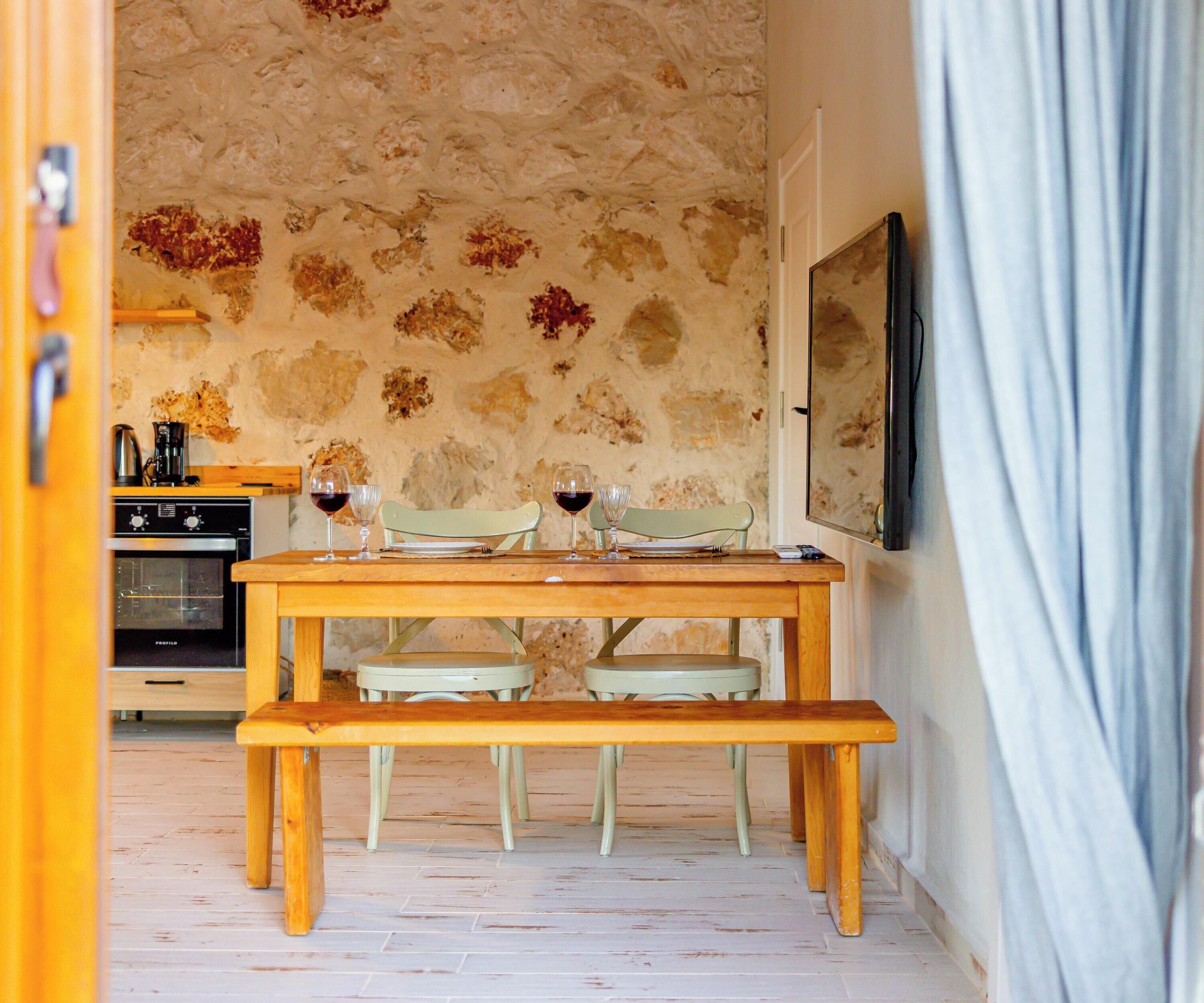 Un comedor rústico con una mesa de madera, dos sillas y un banco. Se observa una estufa antigua al fondo.