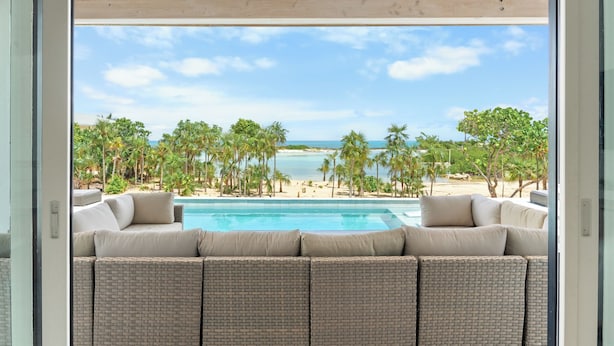 A view from a balcony with a pool, outdoor seating, and a beach with palm trees and the ocean.