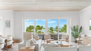 A modern living room with a large window overlooking a beach and palm trees. There are multiple seating areas, including a sofa, chairs, and a dining table.