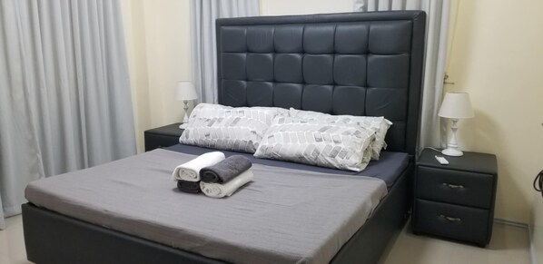 Ein ordentlich bezogenes Bett mit einer grauen Tagesdecke und zwei Kissen, ein Nachttisch mit Lampe sowie ein Beistelltisch mit Schublade.