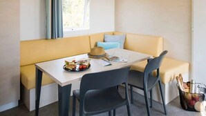 A small, cozy room with a yellow sofa, a wooden table, two chairs, a window with curtains, and a basket of fruits.