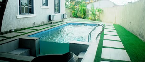 Una piscina con un diseño geométrico de baldosas, junto a un edificio blanco con ventanas y altavoces, rodeada por un muro de concreto y follaje.