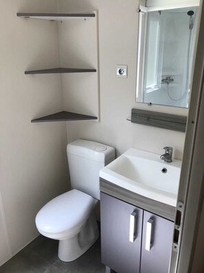Compact bathroom with a toilet, sink, and mirror, featuring storage shelves and grey tile flooring.