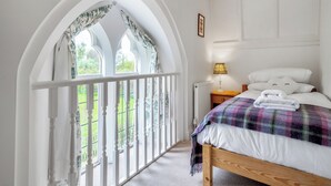 A bedroom with a wooden bed, a nightstand with a lamp, and a view through a window with a white railing. - 4 bedroom accommodation in Hilmarton, near Calne (Hilmarton, near Calne)