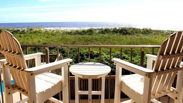 Um deck de madeira com duas cadeiras Adirondack e uma pequena mesa, com vista para uma praia com dunas gramadas e o oceano.