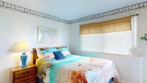 A bedroom with a bed, a nightstand with a lamp, a window with blinds, and a wall-mounted picture.
