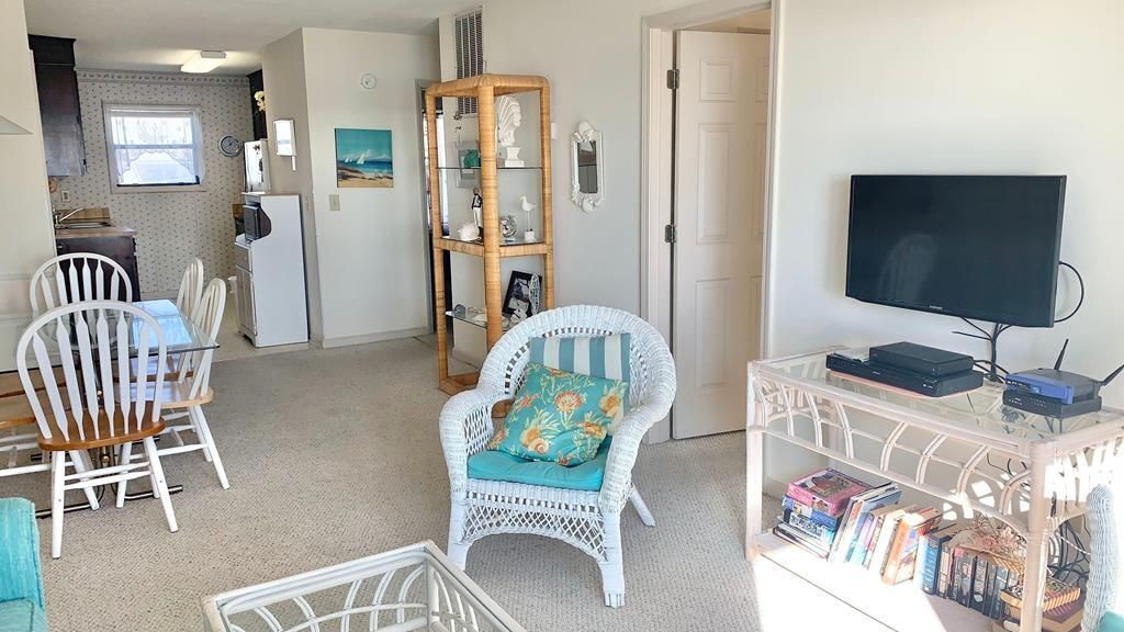 A living room with a white wicker chair, a bookshelf, a television, and a dining area with white chairs.