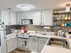 A modern kitchen with white cabinets, a microwave, a stove, and a sink. There is a decorative shelf with various items and a sign that says "BEACH TOWN".