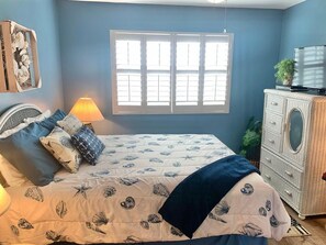 A bedroom with a bed, a dresser, a lamp, and a window with blinds.