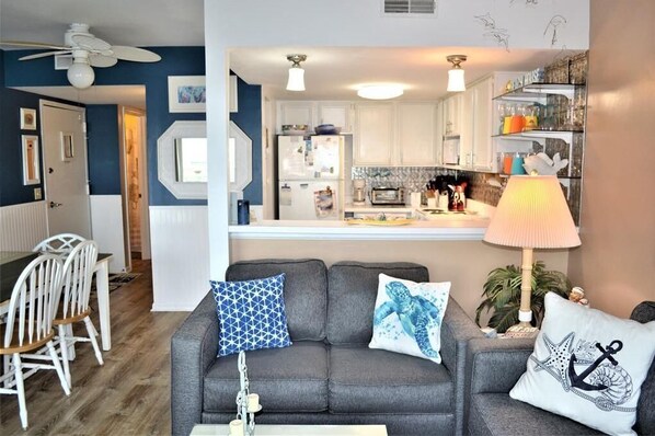 A living room with a grey sofa, white dining table, and chairs, a kitchen with white cabinets and a countertop, and a ceiling fan.