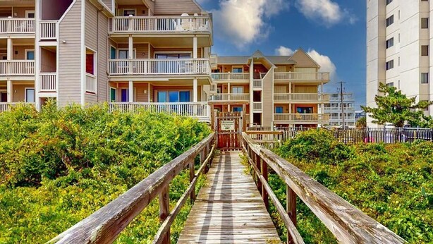 Eine Holzpromenade führt zu einem Strandhotel mit mehreren Balkonen und wolkenlosem Himmel.