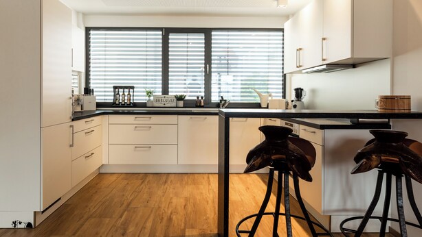 Una cocina moderna con armarios blancos, encimera negra y un amplio ventanal con persianas.
