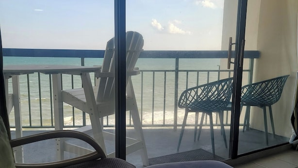 A balcony with a view of the sea, a white chair, a blue cushioned bench, and a green chair.