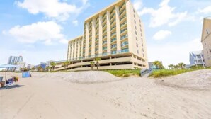A high-rise hotel with multiple balconies, located near a beach with sand dunes and palm trees.