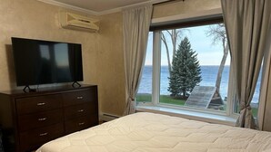 A bedroom with a bed, a TV on a dresser, and a window with a view of the sea and a tree.