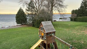 A wooden playhouse with a slide and swing set in a grassy yard with a view of the water.
